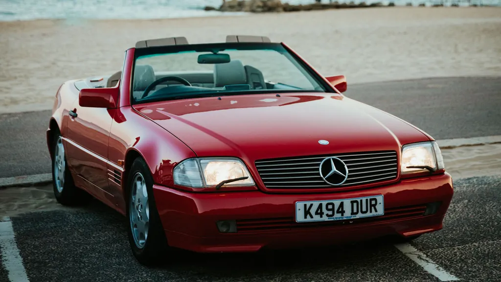 Red Mercedes-Benz SL R129 convertible parked near the beach with roof down, front three-quarter view